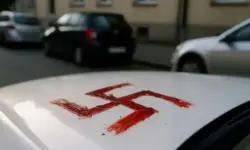 Nahaufnahme eines weißen Autodachs mit roter Flüssigkeit in Form eines Hakenkreuzes, im Hintergrund unscharf geparkte Autos und Häuser.