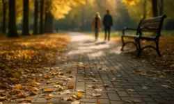 Herbstlicher Parkweg mit buntem Laub, Parkbank und zwei unscharfen Personen im Hintergrund bei warmem Sonnenlicht.