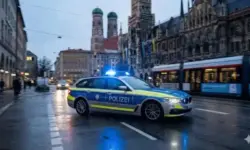 Ein silber-blauer BMW-Kombi der Polizei Bayern fährt mit eingeschaltetem Blaulicht auf einer nassen Straße bei Dämmerung. Im Hintergrund sind die Zwillingstürme der Frauenkirche und das Neue Rathaus in München zu erkennen.