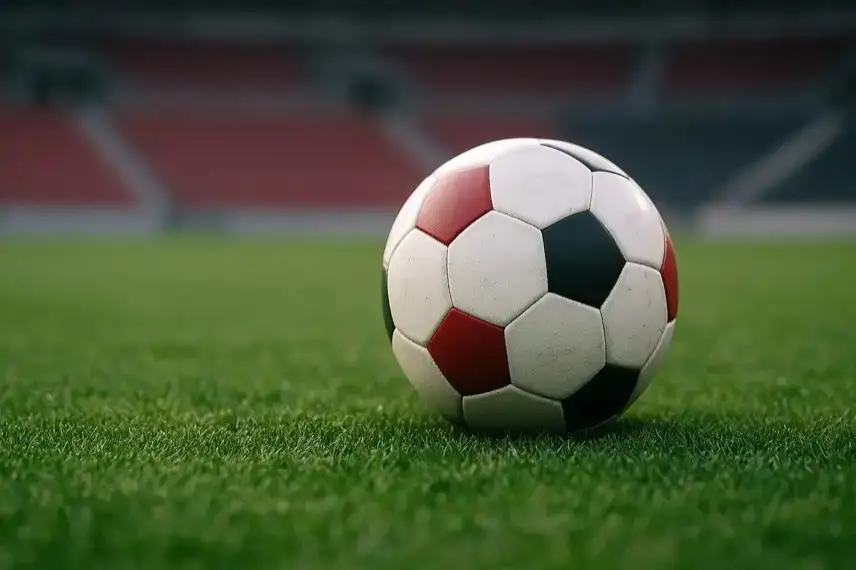 Leerer Fußballplatz mit einem einzelnen Spielball auf grünem Rasen, unscharfe Stadiontribüne im Hintergrund, aufgenommen bei natürlichem Licht.