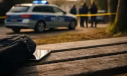 Parkbank mit schwarzer Tasche und Smartphone im Vordergrund, im Hintergrund ein Polizeifahrzeug mit eingeschaltetem Blaulicht und Absperrband.