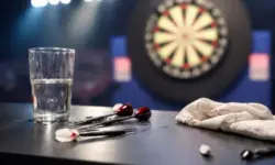 Leere Darts-Bühne mit Dartboard im Hintergrund und beschädigtem Tisch mit umgekipptem Wasserglas im Vordergrund.