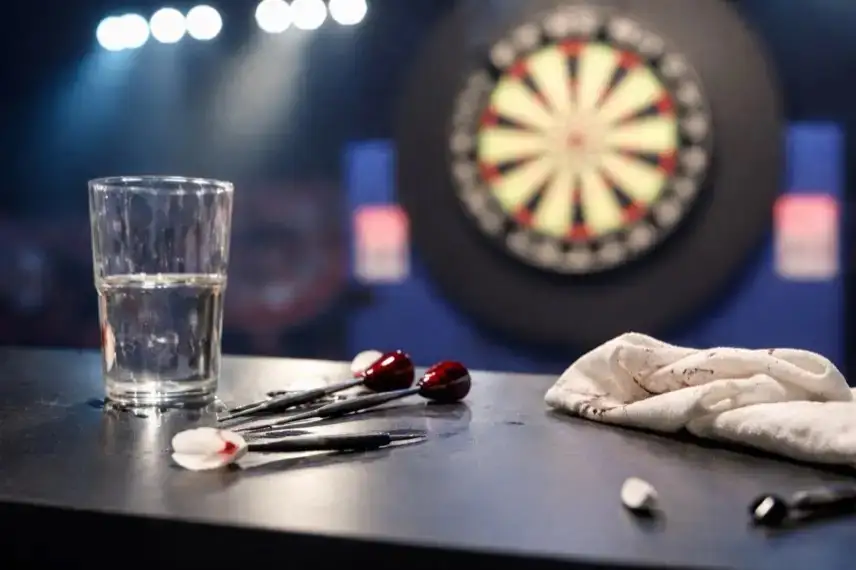 Leere Darts-Bühne mit Dartboard im Hintergrund und beschädigtem Tisch mit umgekipptem Wasserglas im Vordergrund.
