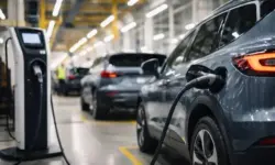 Mehrere neutrale Elektrofahrzeuge auf einem Parkplatz mit Ladesäulen in einem modernen Industrieumfeld bei natürlichem Licht.