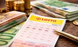 Lottoschein auf einem Holztisch, daneben gestapelte Euro-Banknoten, Münzen und ein Kugelschreiber in hellem Licht.