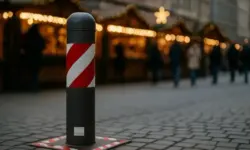 Ein schwarz-grauer Poller mit rot-weißer Markierung auf Kopfsteinpflaster, im Hintergrund unscharfe Weihnachtsmarktstände mit Lichterketten.