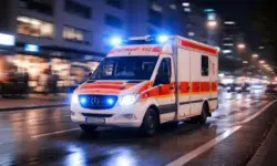 Krankenwagen mit Blaulicht fährt nachts durch eine Stadt, nasse Straße, unscharfe Gebäude im Hintergrund.
