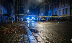 bgesperrte Straße vor einem Hotel mit mehreren Polizeifahrzeugen und Blaulicht bei winterlichem Tageslicht, rot-weißes Absperrband im Vordergrund, keine Menschen sichtbar.
