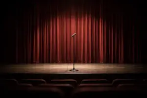 Leere Theaterbühne mit geschlossenem Vorhang und einem einzelnen Mikrofonständer im Vordergrund, neutral beleuchtet, ohne Personen.