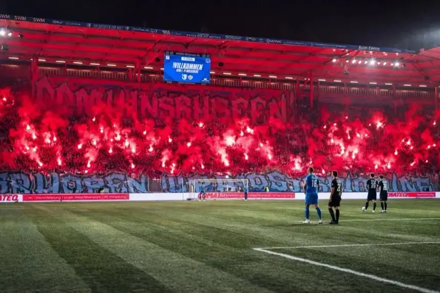Polizeifahrzeuge und Absperrgitter vor einem Stadion bei Nacht, Rauchschwaden im Hintergrund nach einem Fußballspiel