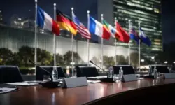 Mehrere Flaggen vor dem UN-Gebäude bei Nacht, davor ein leerer Konferenztisch mit Mikrofonen als Symbol für internationale Diplomatie