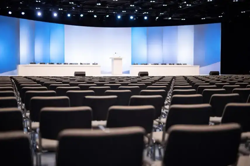 Leere Parteitagshalle mit Reihen von Stühlen und einem Rednerpult, hell ausgeleuchtet, neutrales politisches Veranstaltungsambiente.