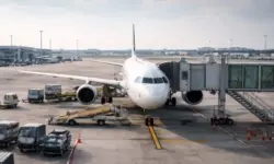 Verkehrsflugzeug steht an einem Flughafen-Gate, umgeben von Bodenfahrzeugen und Fluggastbrücke bei Tageslicht.